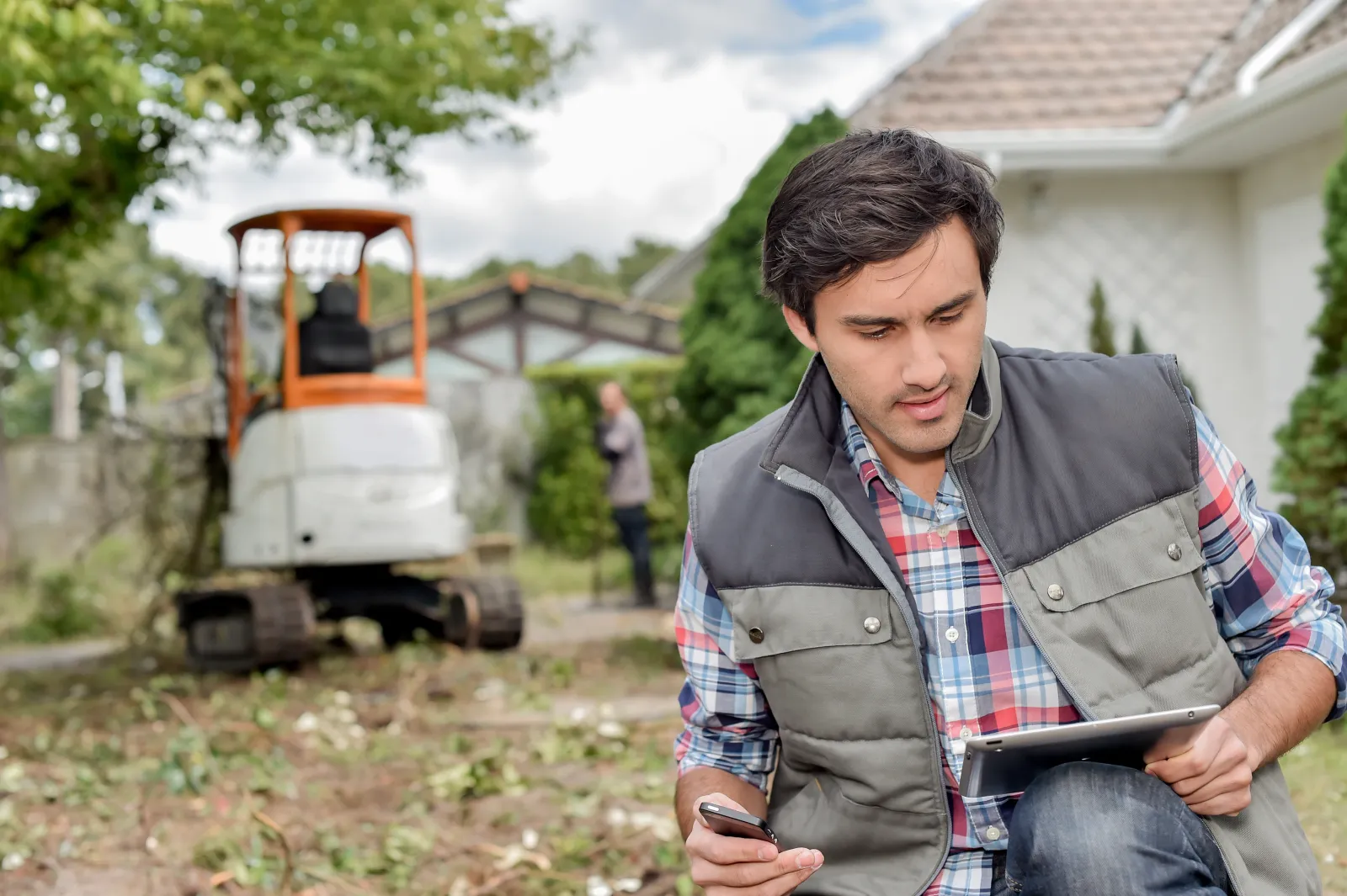 Landscaping contractor outside using tablet, digger in background